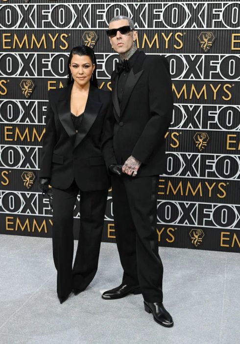 Kourtney and Travis, both in sleek suits, strike a pose on the red carpet with an Emmys backdrop. Kourtney's height reaches just up to his earlobe.