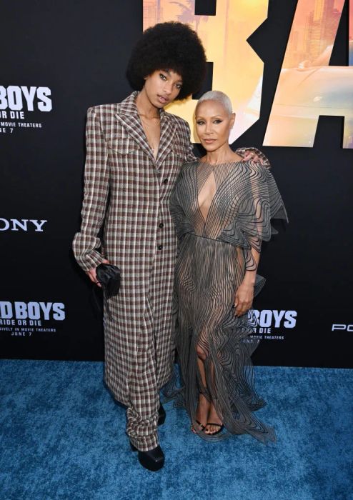 Willow and Jada grace the red carpet: Willow in a plaid suit holding a clutch, while Jada wears a sheer dress with a geometric pattern. Both are in heels, with Jada reaching up to Willow's jawline.