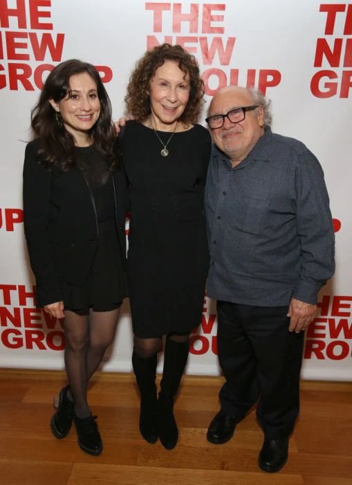 The family is photographed together at a New Group event. The women wear dresses, with one sporting boots, while Danny wears glasses and a casual shirt. Danny's height reaches just up to Rhea's eyes.