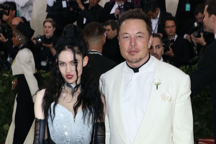 Grimes and Elon Musk at a red carpet event: one dressed in a gothic-style outfit, the other in a white suit paired with a black shirt.