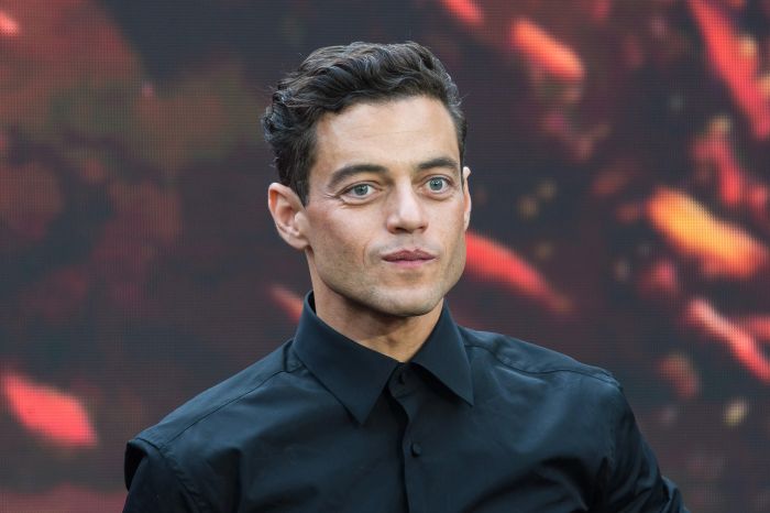 Rami Malek, dressed in a black shirt, stands against a blurred background with a faint smile.