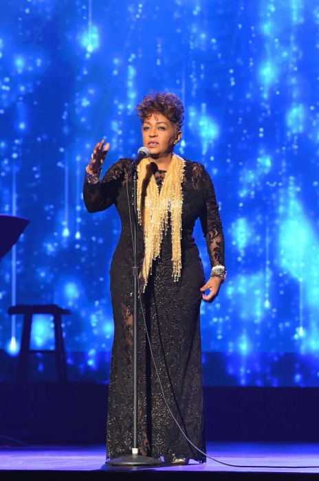 Anita, wearing an elegant, long lace gown and a beaded necklace, stands on stage holding a microphone, with a starry backdrop behind her.