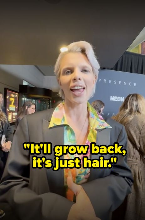 A person stands at a media event, wearing a black blazer over a colorful shirt, in front of a backdrop that says 'NEON,' with people gathered in the background.