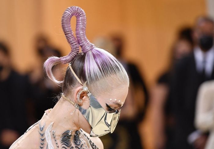 Grimes sporting a detailed metal face mask and a purple, spiraled hairpiece on the red carpet, dressed in a gown adorned with striking tattoos.