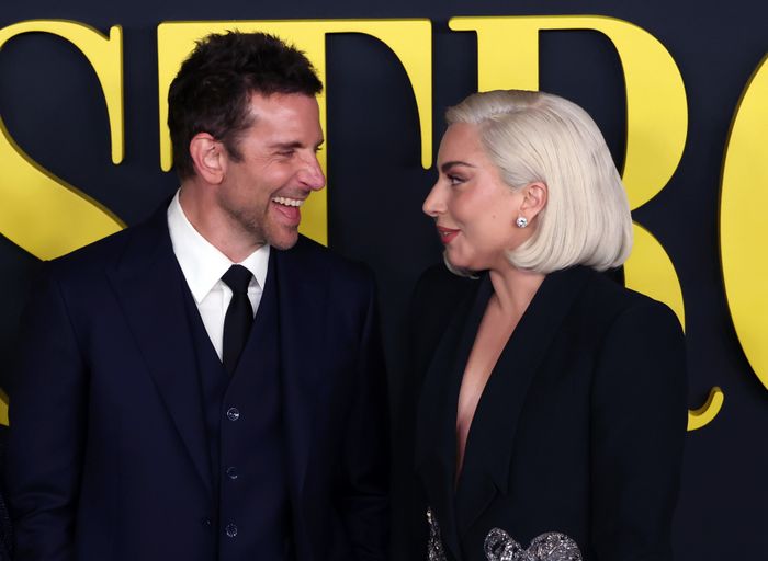 A pair of people exchanging smiles on the red carpet, both dressed in elegant, dark-colored outfits, with a backdrop featuring large yellow text.