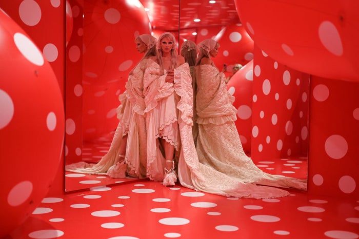 A person stands elegantly in a detailed lace gown with a sweeping train, set against a vibrant red room dotted with white polka dots and framed by mirrors.