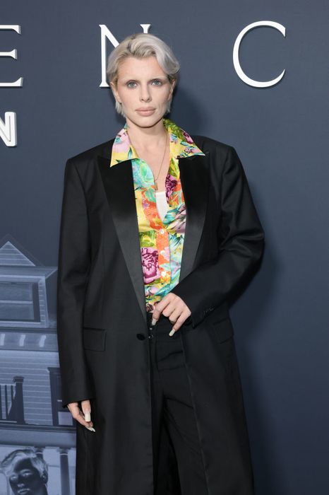 A person stands on the red carpet, wearing a floral shirt paired with a black blazer, gazing directly at the camera with confidence.