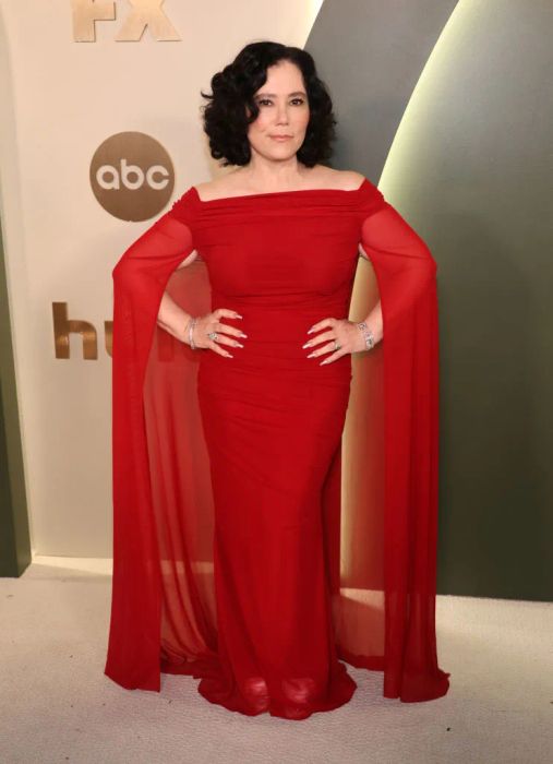 Alex poses in a flowing, off-the-shoulder gown at a red carpet event, with her hands placed confidently on her hips.