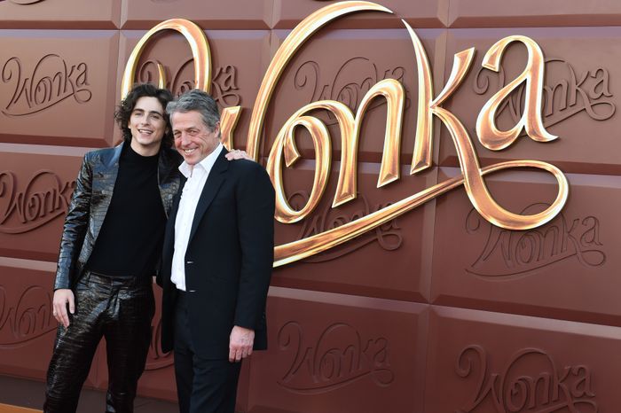 Two individuals strike a pose at the 'Wonka' premiere. One is dressed in a shiny suit, while the other wears a classic suit. They stand against a backdrop featuring the 'Wonka' text.
