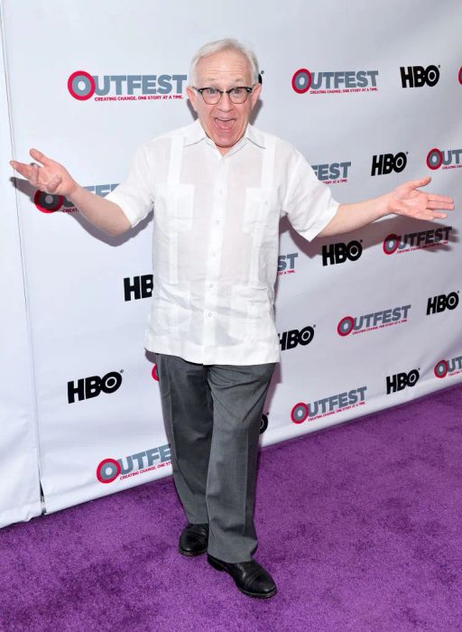 Leslie is pictured on a red carpet at an Outfest event, dressed in a short-sleeve shirt and pants, with his arms wide open in a gesture of enthusiasm.