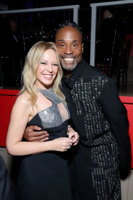 Kylie dons a dress with metallic accents, while Billy, with an arm around her, sports a jacket adorned with text patterns. She reaches up to his eyes.