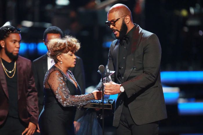 Anita, dressed in an elegant gown with sheer sleeves, accepts a star-shaped award from Tyler, who is in a suit. Other people can be seen behind them on stage. She doesn't quite reach his shoulder.