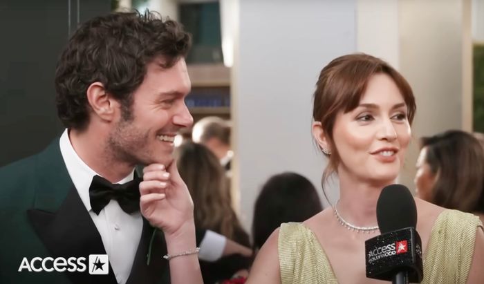 At a glamorous red carpet event, a man in a classic tuxedo smiles warmly while a woman in an elegant sleeveless gown addresses the crowd with a microphone. The Access Hollywood logo is visible in the background.