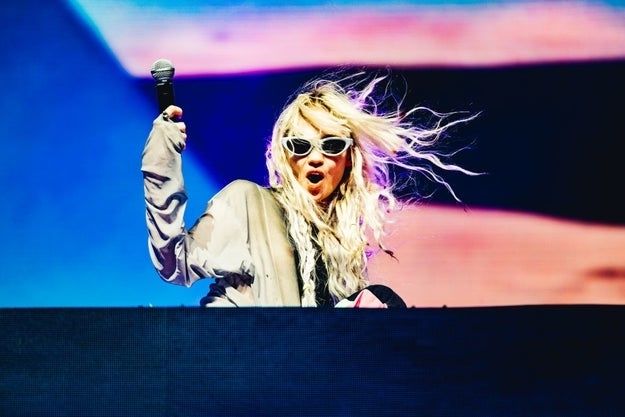 Grimes energetically performing on stage, wearing a loose outfit and signature sunglasses, holding a microphone with her hair flowing in motion.