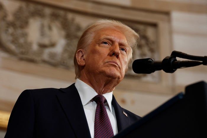 Donald Trump addresses the audience at a podium, his expression serious, dressed in a dark suit and tie.