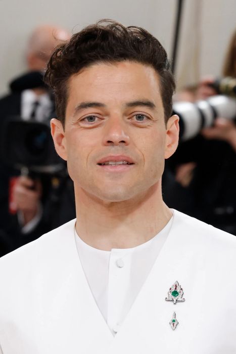 Rami Malek, wearing a formal white outfit with decorative pins, stands at an event surrounded by photographers.
