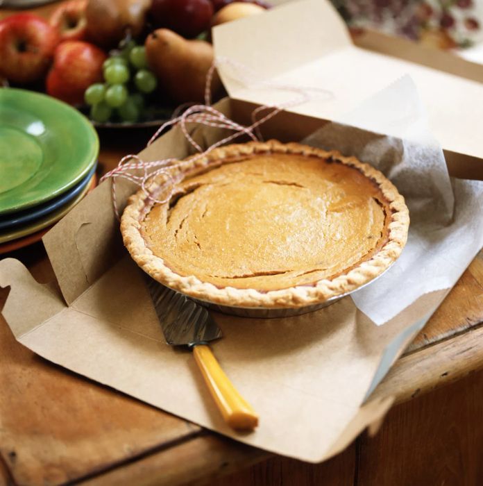 A slice of pumpkin pie with a serving knife placed in an open box, accompanied by a stack of colorful plates and a basket of fresh fruit.