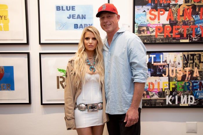 The couple can be seen standing in front of some artwork; the woman is dressed in a white gown with turquoise jewelry, while the man sports a red cap and blue shirt.