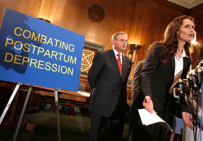 Brooke speaks at a podium next to a sign reading 'Combating Postpartum Depression', with a man in a suit standing behind her.