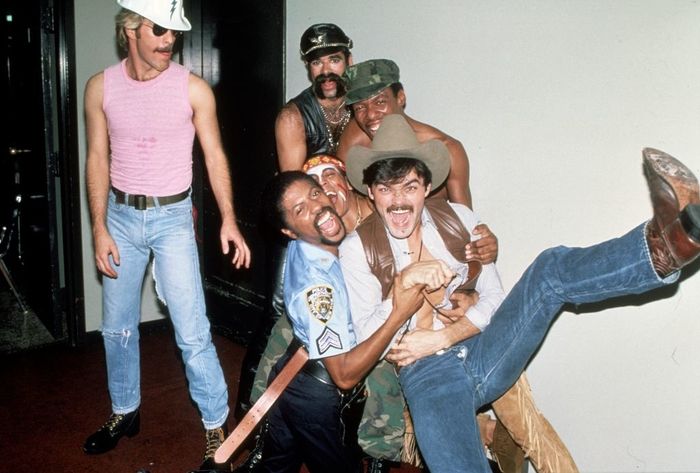 A group of five men in themed costumes, each resembling traditional uniforms, strike playful poses. One sports a construction helmet, another dons a police uniform, and the rest are similarly attired.