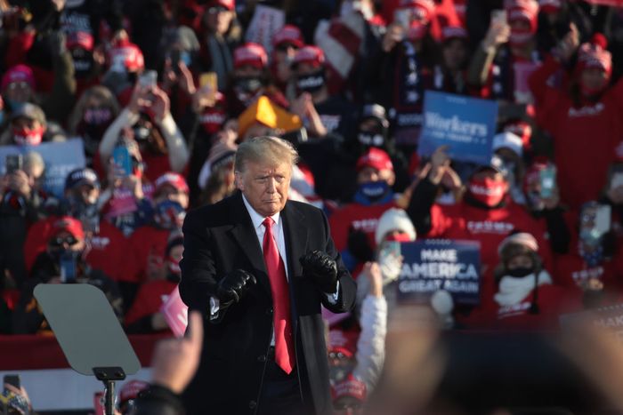 Trump, dressed in a suit and red tie, stands outdoors at a rally, gesturing as a crowd cheers with signs in hand behind him.