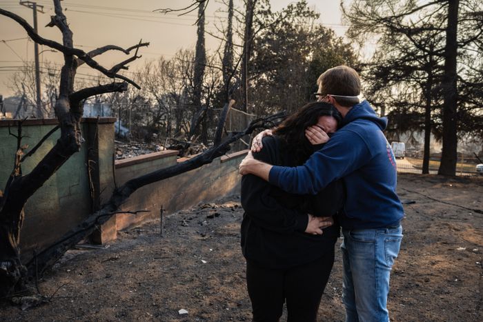 Two people share a heartfelt embrace amidst a devastated landscape, with charred trees and scattered debris, capturing the raw emotion of loss and disaster's aftermath.