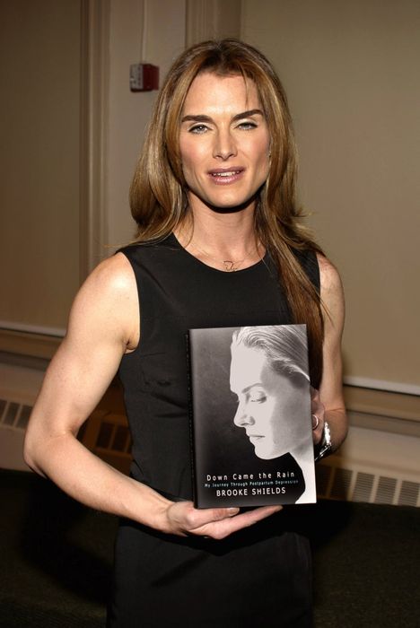 Brooke smiles while holding her book titled 'Down Came the Rain', wearing a sleeveless dress at an event.