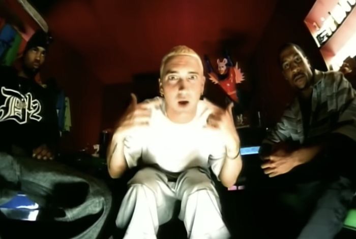 Three people in a dim room, one standing in the center, making gestures towards the camera, dressed in casual streetwear.