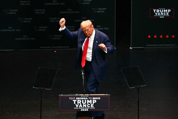 A person in a suit gestures on stage with 'Trump Vance 2024' signage and text prompts urging Georgia voters in the background.