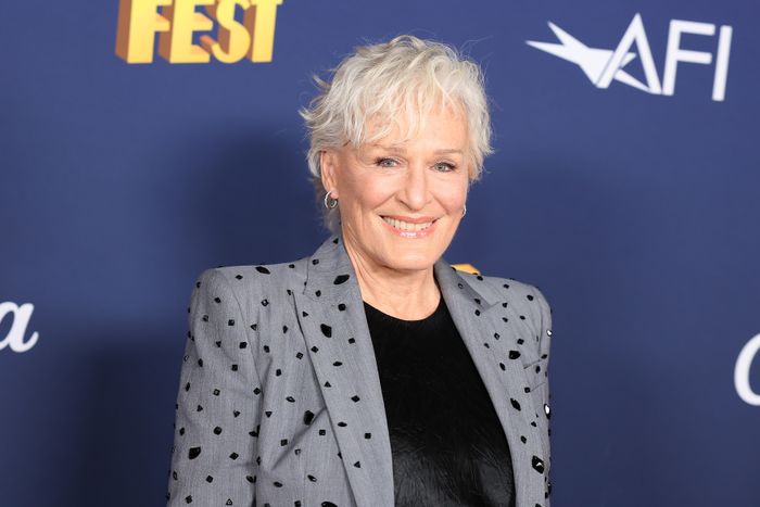Glenn Close flashes a bright smile while wearing a patterned blazer and sporting short hair, standing against a backdrop full of logos and text at an event.