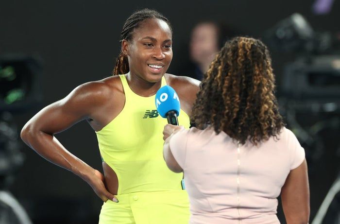 Coco smiles brightly during a post-match interview on the court, her hands placed confidently on her hips as she dons a sleeveless athletic suit.