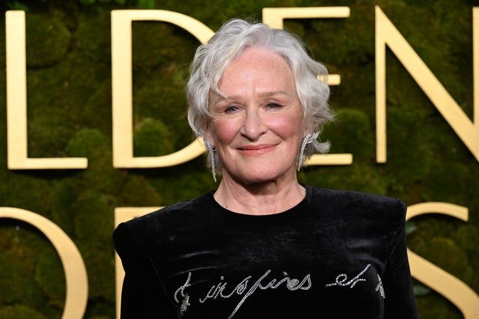 A radiant Glenn Close beams on the red carpet, dressed in a velvet gown adorned with intricate embroidered script. Behind her, large golden letters shine.