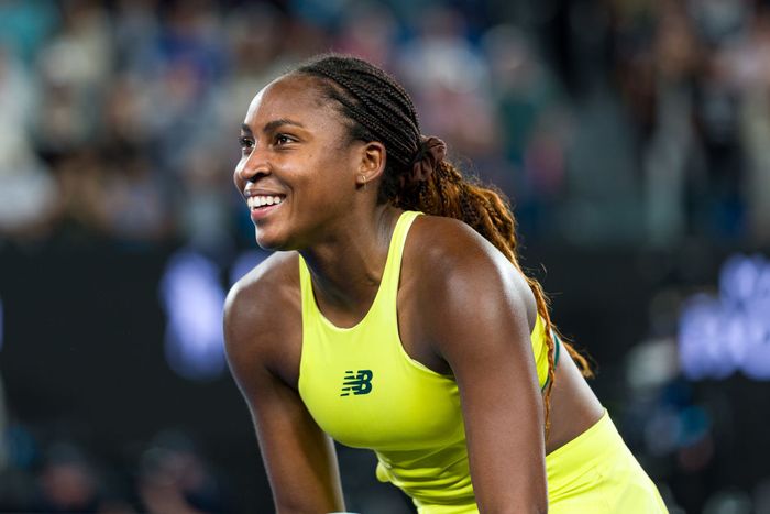 An athlete flashes a bright smile while leaning forward on a tennis court, dressed in a sporty tank top and shorts.