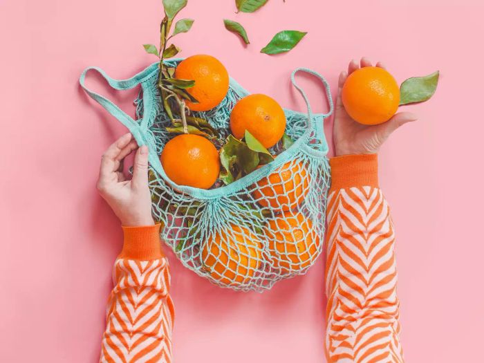 A woman wearing an orange striped sweatshirt holds oranges with green leaves in a blue eco-friendly mesh shopping bag against a pink background.