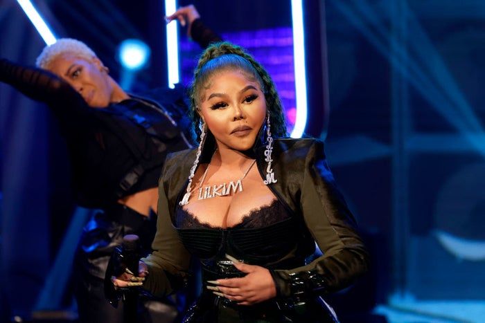 Lil' Kim takes the stage in a fashionable outfit, accessorized with bold statement pieces, microphone in hand, with a background dancer adding to the energetic atmosphere of the performance.