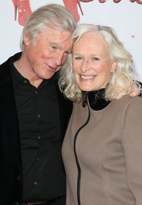 David Shaw and Glenn Close share a warm smile, with one arm around the other, dressed in dark and neutral tones, posing in front of a backdrop.