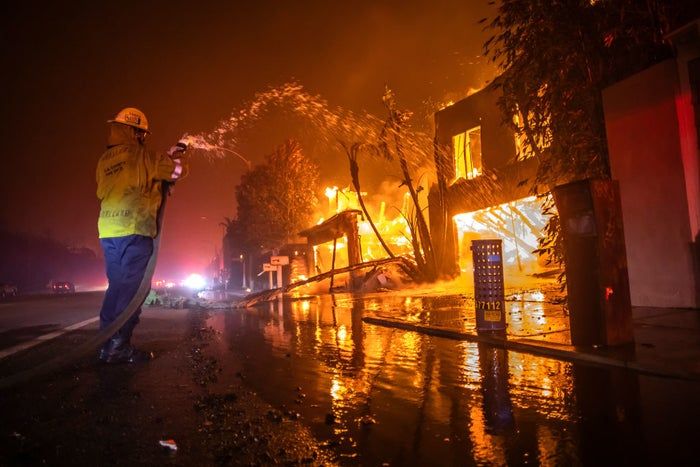 A firefighter in full gear sprays water on a blazing house during a night-time firefighting effort. The emergency response continues.