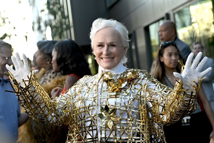 Glenn Close, dressed in a detailed gold wire outfit with floral embellishments, smiles and waves at an outdoor event, with people in the background.