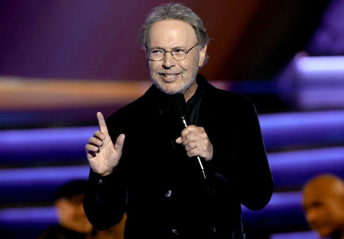 A person in a deep velvet suit holds a microphone and gestures while speaking to a crowd of listeners.