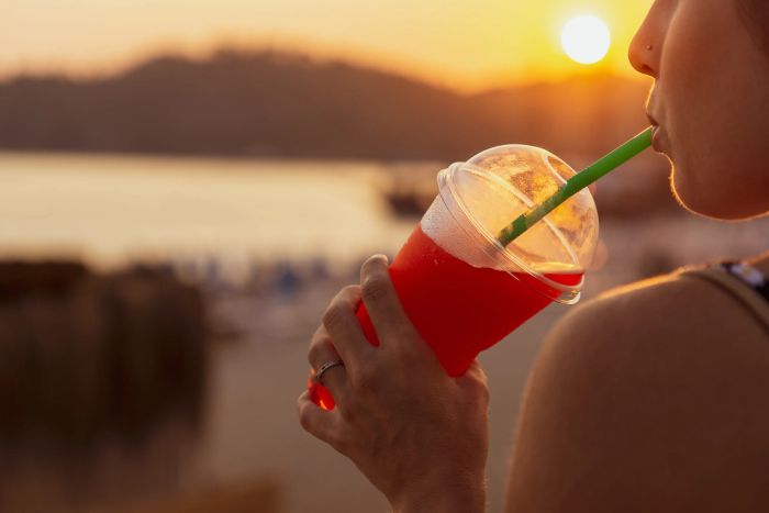 A woman sipping an icee while the sun sets over the beach in the background.