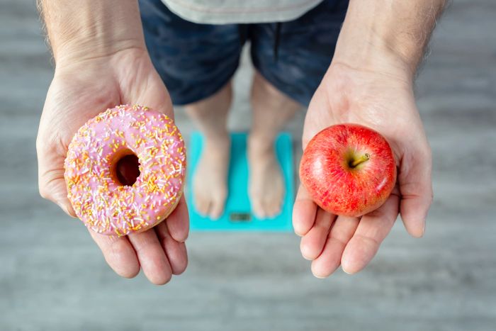 A person stands on a scale, holding a donut in one hand and an apple in the other, symbolizing a choice between unhealthy and healthy food options.