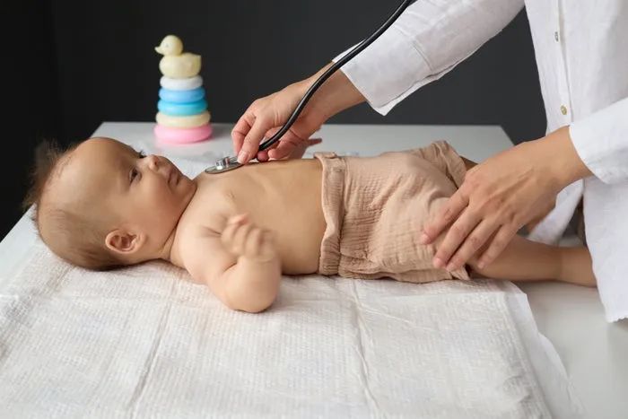 A doctor examines a baby lying on a table with a stethoscope, while a toy tower can be seen in the background.