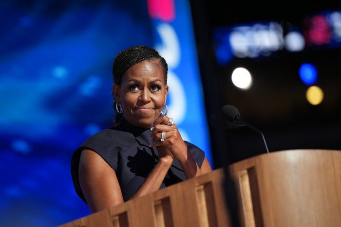 Michelle Obama, dressed in a sophisticated sleeveless gown, addresses an audience at an event, holding a microphone.