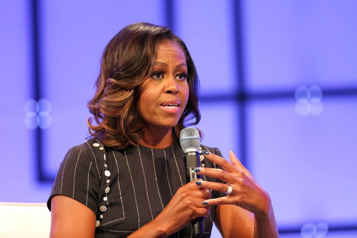 Michelle Obama, dressed in a pinstripe dress, speaks on stage, holding a microphone and gesturing animatedly with one hand.