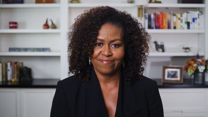 Michelle Obama, with curly hair and dressed in a blazer, stands confidently in front of a bookshelf.