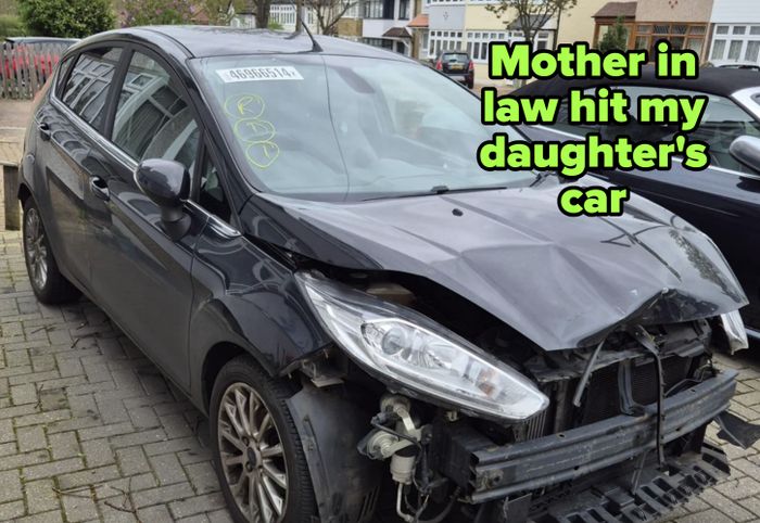 A damaged black car with a front-end collision, missing its front bumper and headlight, parked on a residential street. QR codes visible on the windshield.
