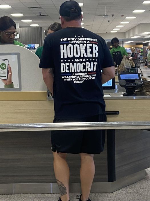 A person with a shirt featuring bold text at a store checkout counter. Employees in green uniforms assist.
