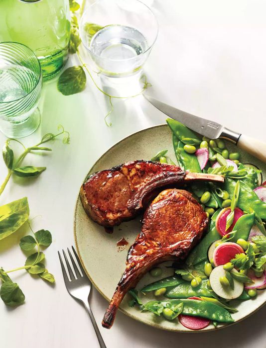 Two Five-Spice Lamb Chops served with a snow pea and radish salad, plated with metal utensils and accompanied by glasses of water.