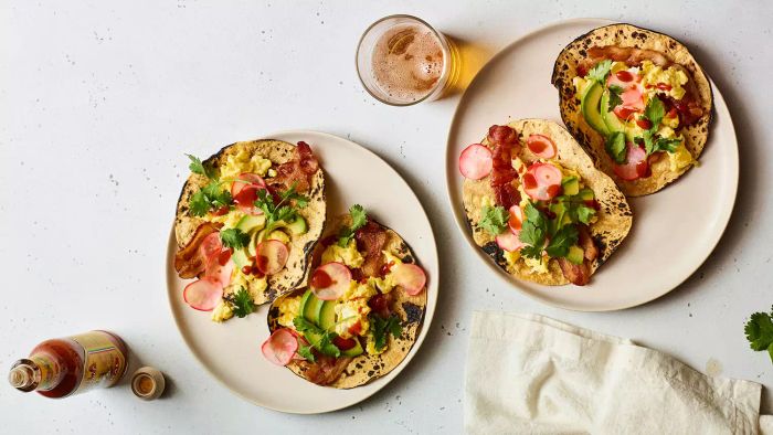 Four crispy bacon and egg tacos, topped with radishes and avocado, served in pairs of two on two separate plates.