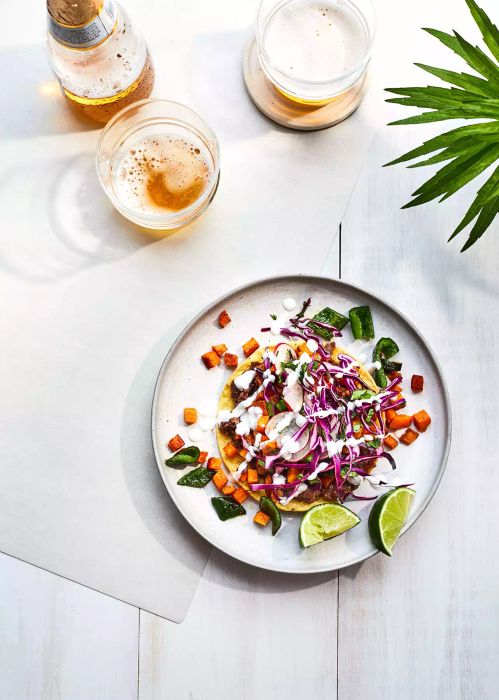 Black Bean Tostadas with Sweet Potato and Poblanos served on a plate, accompanied by lime wedges and two glasses of beer, set on a table.